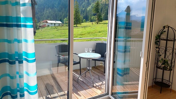 Une pièce dotée d’une porte-fenêtre coulissante donnant sur un balcon avec une table et des chaises, d’où l’on peut admirer un paysage verdoyant avec des arbres et une maison, ainsi qu’un présentoir métallique pour plantes agrémenté de fleurs.