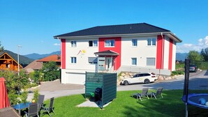 Un bâtiment à deux étages avec une façade rouge et blanche, un espace extérieur couvert avec des sièges, et une voiture blanche garée devant.