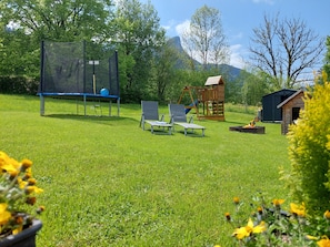 Ein grünes Außenbereich mit Trampolin, zwei Liegestühlen, einer Holzschaukel und einem kleinen Schuppen.