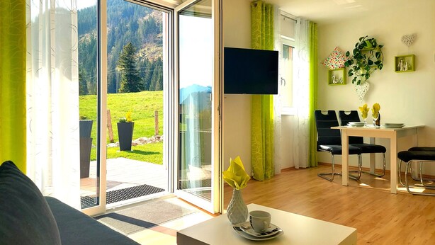 Un salon comprenant un canapé, une table avec un vase et une tasse, un téléviseur fixé au mur, et un coin repas avec des chaises et une table.