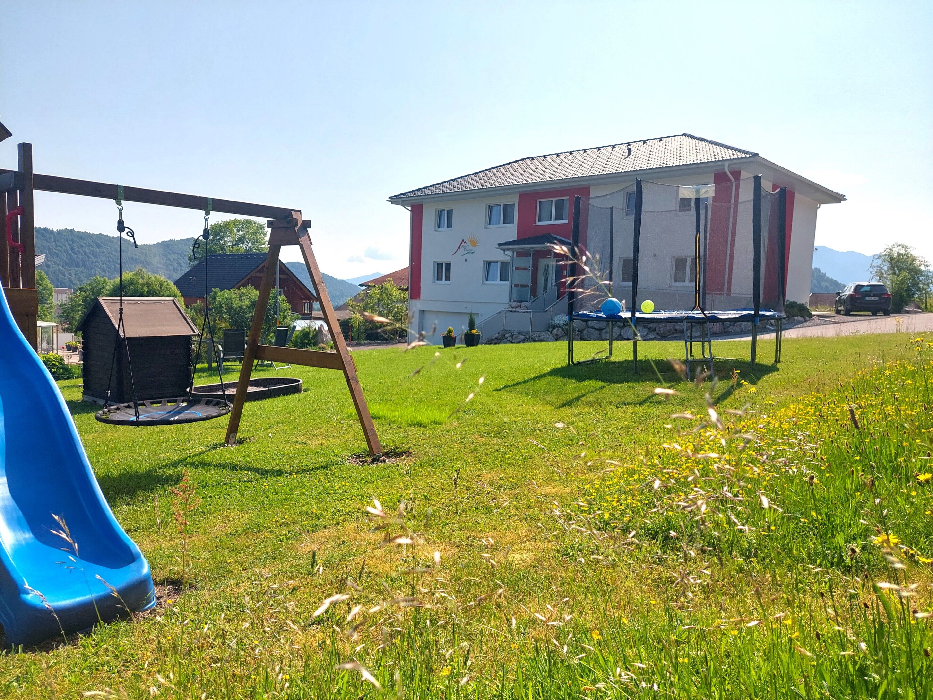 Un parque infantil con un tobogán azul y columpios, un trampolín y un edificio de fachada roja y blanca.