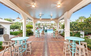 A covered outdoor area with tables and chairs, a pool in the background, and a building with balconies. - Sea Blessings- A Beautiful 4 Bedroom Home Steps from Seacrest Beach! (Inlet Beach)