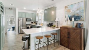 A modern kitchen with light green cabinets, stainless steel appliances, a white island with stools, and a wooden cabinet with decorative items.