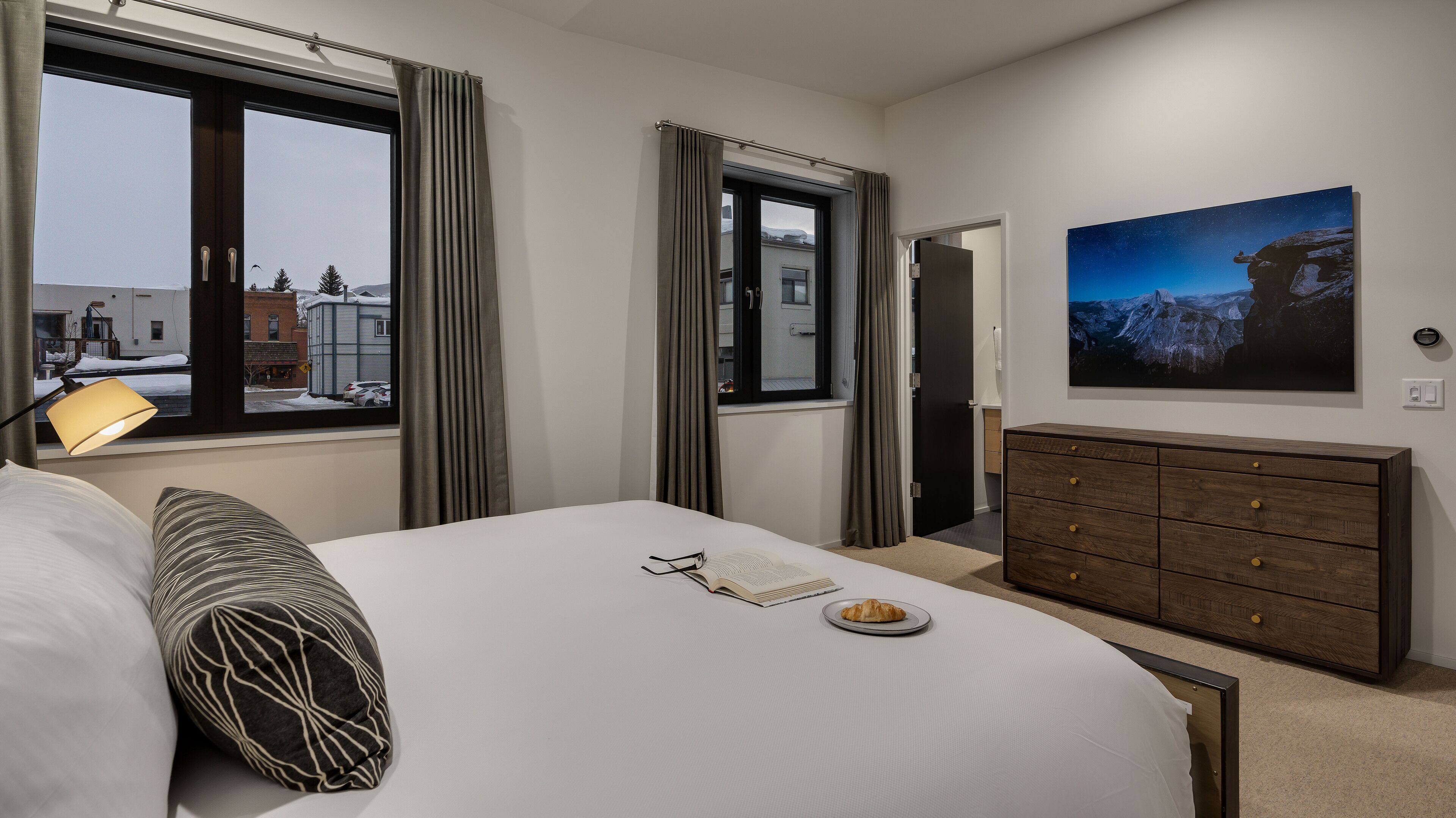 A hotel room with a bed, a dresser, a window with curtains, a lamp, and a painting on the wall.