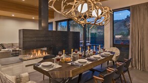 Un espace repas moderne avec une longue table dressée pour six personnes, une cheminée et une grande fenêtre donnant sur l’extérieur.
