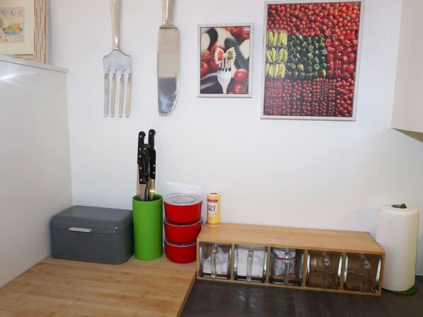 Una cucina con un piano di lavoro in legno, una scatola portaoggetti grigia, un contenitore verde e un contenitore rosso. Si vedono coltelli in un portacoltelli, una lattina di sugo e una rastrelliera in legno con utensili.
