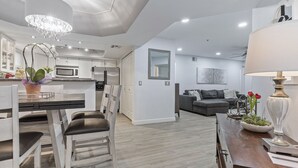 Una cocina moderna con armarios blancos, un comedor con mesa y sillas de madera, y una sala de estar con sofá gris y una lámpara.