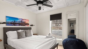 A bedroom with a bed, a ceiling fan, a cityscape painting, a window with a view, and a dark blue bean bag chair.