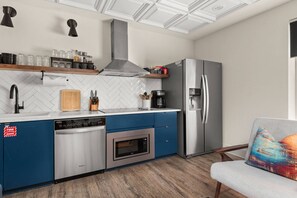 A compact kitchen with a dishwasher, oven, and refrigerator, featuring a herringbone tile backsplash and a wooden cutting board.