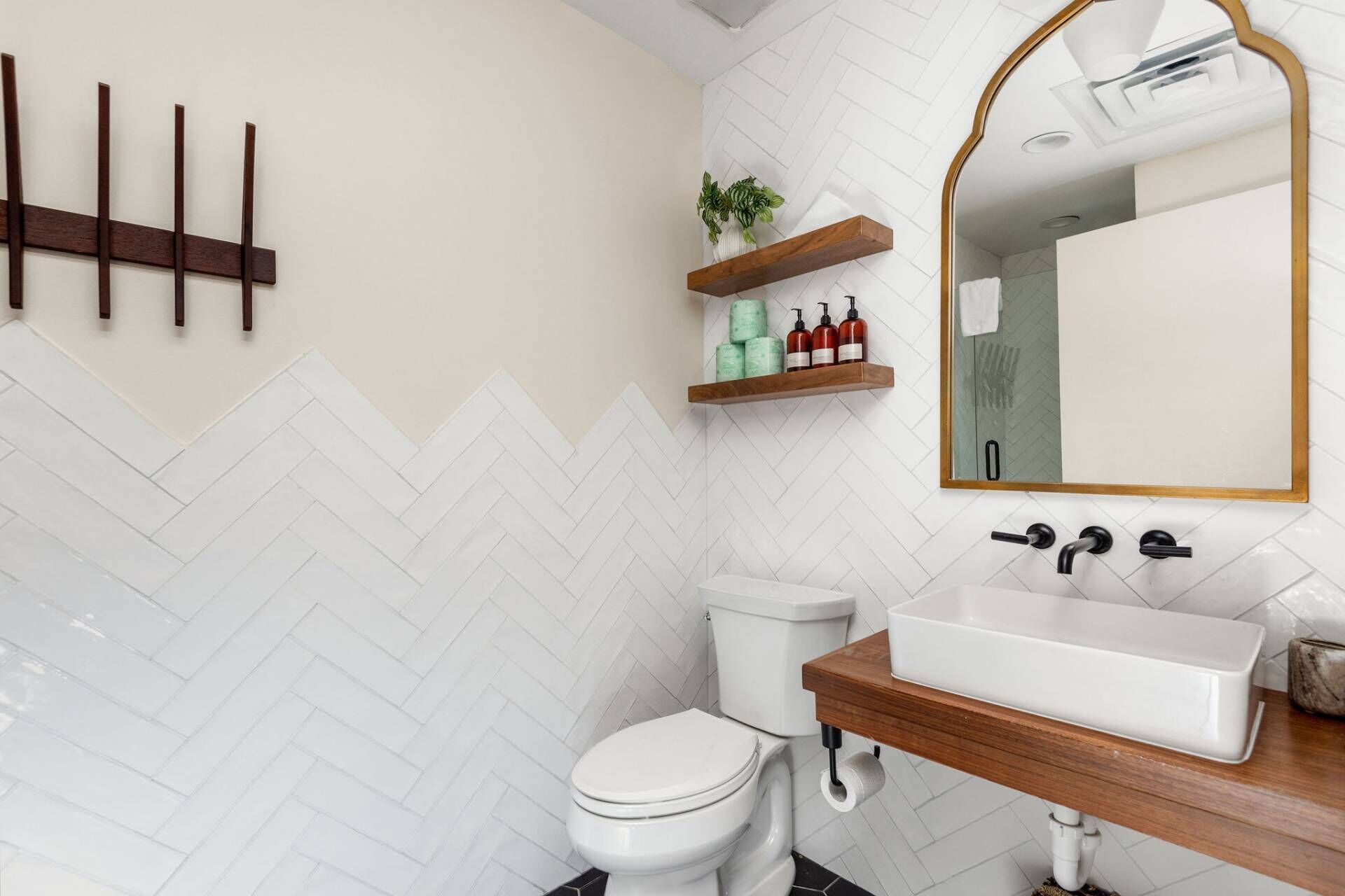 A bathroom with a white toilet, a wooden vanity with a white sink, and a mirror with a gold frame. There are wooden shelves above the sink holding toiletries.
