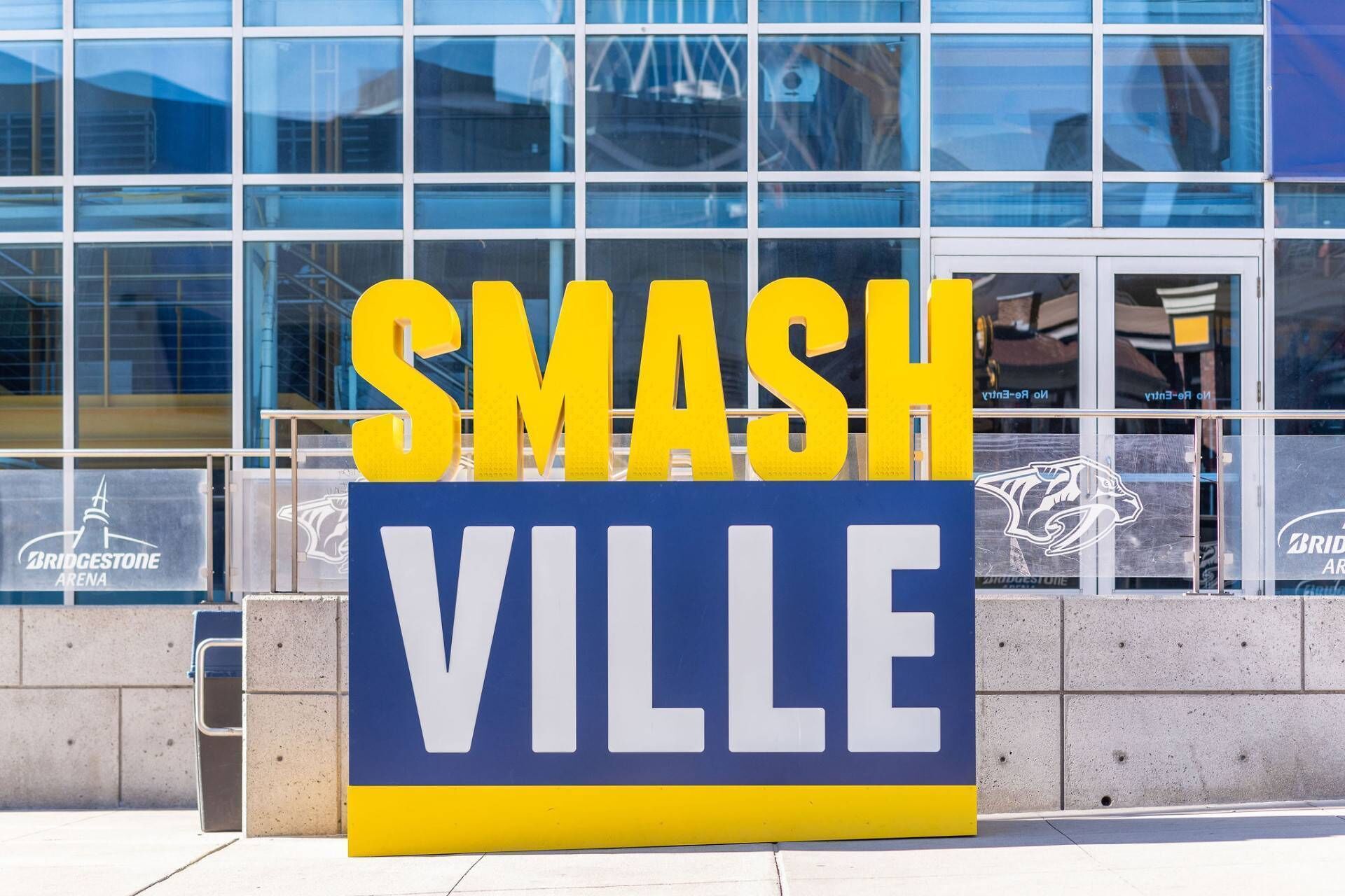 A large sign with the words "SMASH VILLE" in bold yellow letters on a blue background, placed in front of a modern building with glass windows.