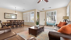 A spacious living room with a dining area, a ceiling fan, and large windows with shutters. - 4-BR Home with a Private Heated Pool (Litchfield Park)