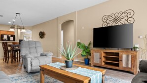 A living room with a grey sofa, a wooden coffee table, a TV on a wooden stand, and a dining area with wooden chairs and a table. - 4-BR Home with a Private Heated Pool (Litchfield Park)