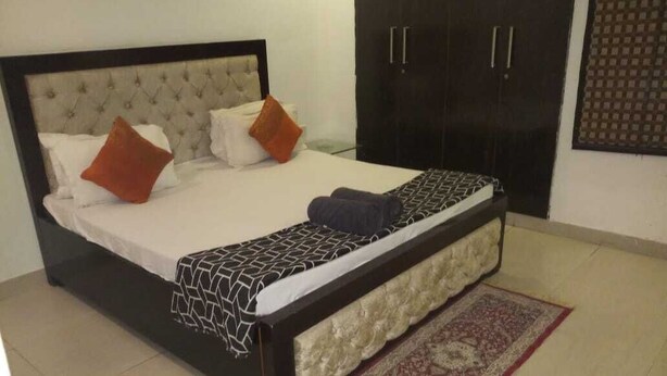 Un lit bien fait, recouvert d’une couvre-lit à motifs, avec deux oreillers et une serviette pliée. On y trouve également une armoire sombre à deux portes et une décoration murale à motifs.