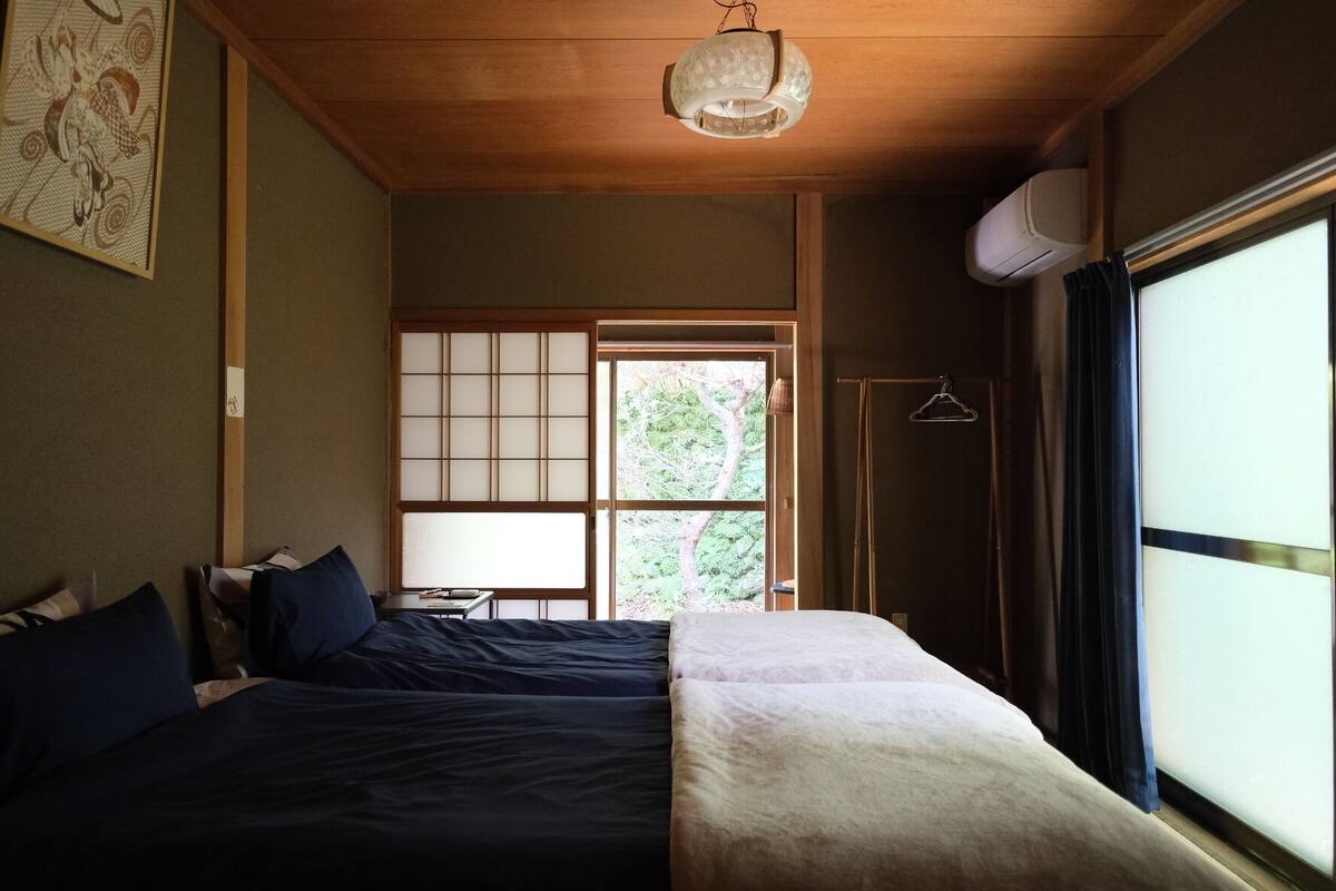 Una habitación con cama, lámpara colgante, ventana con vistas a zonas verdes y aire acondicionado instalado en la pared.