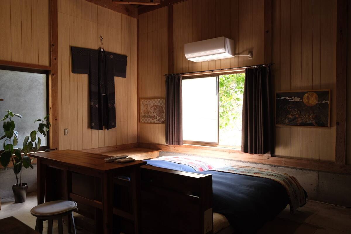 Una habitación con paredes de madera, una cama, un escritorio con una silla, una planta en maceta y una ventana con cortinas.