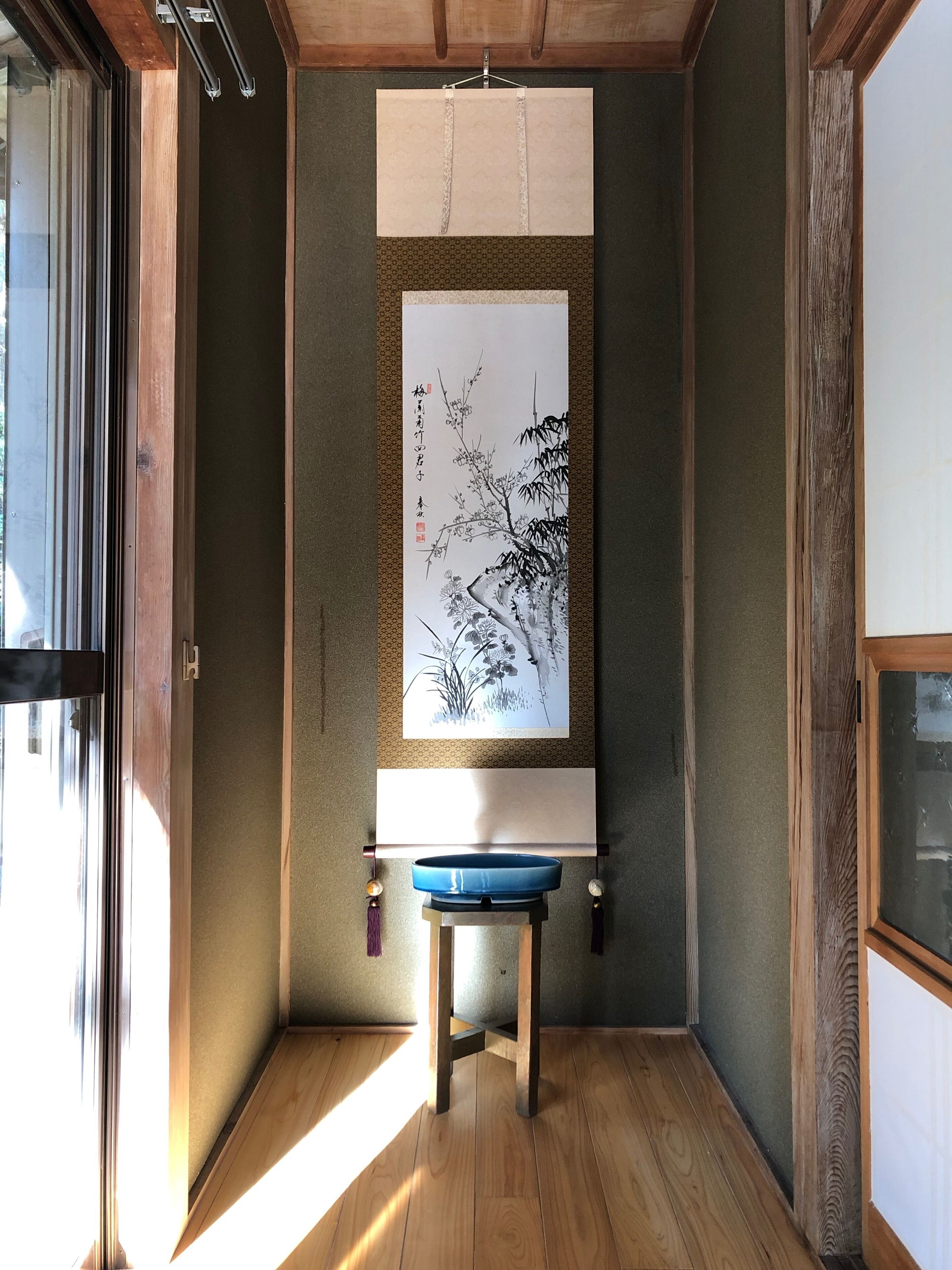 A room with a wooden floor, a blue stool, a framed artwork on the wall, and a window allowing natural light.