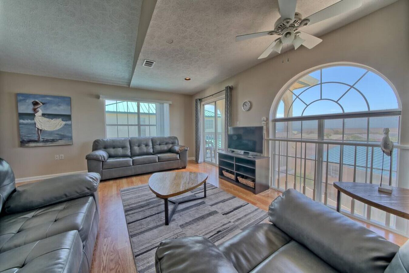 A spacious living room with a grey leather sofa, a coffee table, and a TV stand. There is a large window offering a view of the outdoors.