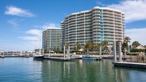 Un hôtel situé au bord de l’eau, doté de nombreux balcons, un port de plaisance avec des bateaux et des palmiers.