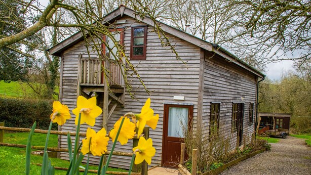 A rustic wooden house with a wooden porch, a small wooden structure to the right, and a gravel path leading to the house. - 3-bed Barn Eco-lodge on the Tarka Trail (Torrington)