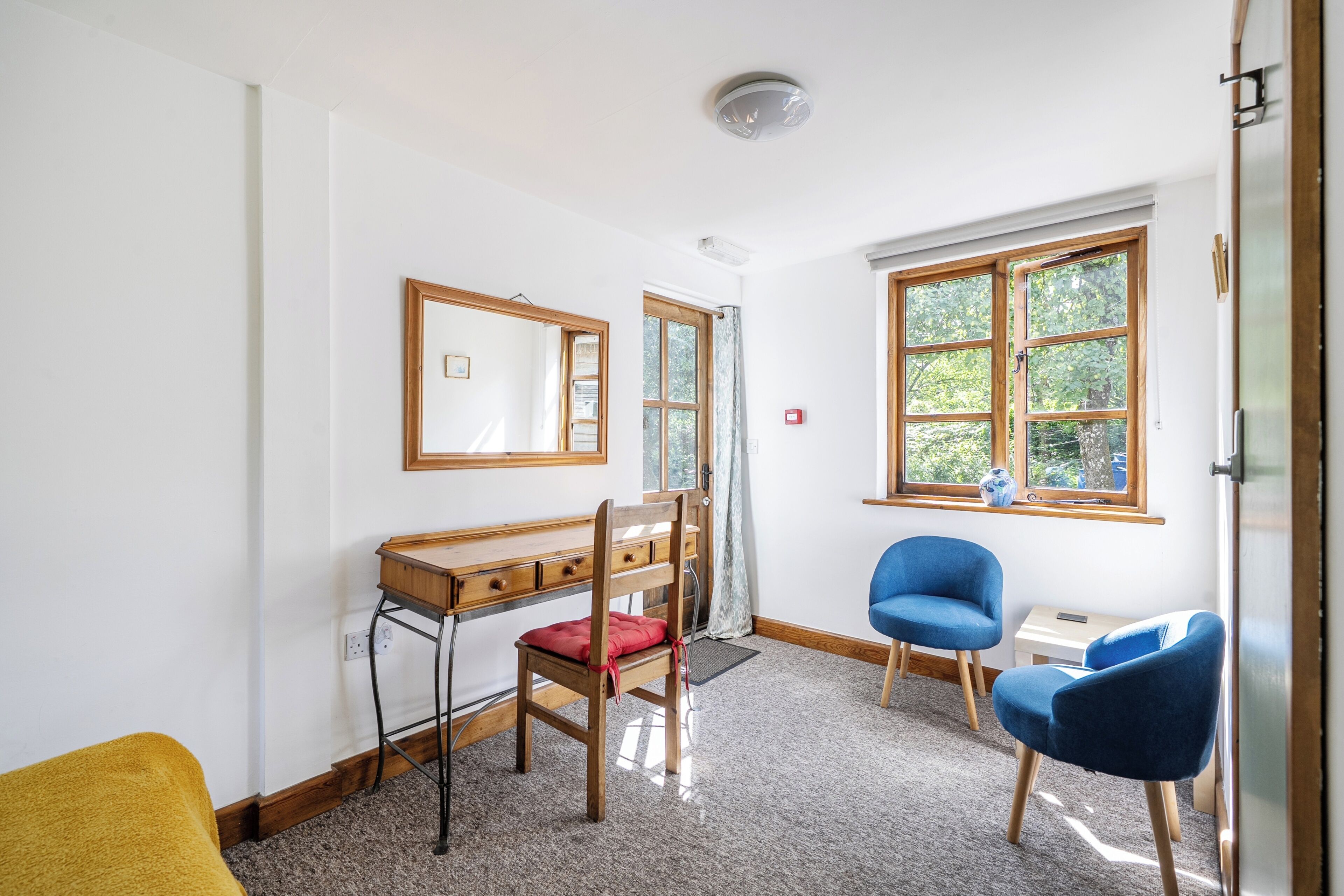 Una habitación con escritorio, dos sillas, ventana con vista a árboles y una lámpara de techo.