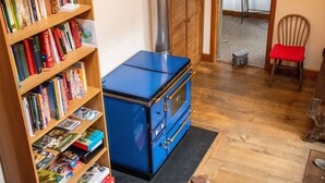 A room with a blue stove, wooden bookshelves, a wooden chair, and a small table. - 3-bed Barn Eco-lodge on the Tarka Trail (Torrington)