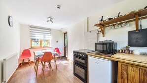 A compact kitchen with a stove, microwave, and a small refrigerator. There is a dining table with chairs and a window with blinds. - 3-bed Barn Eco-lodge on the Tarka Trail (Torrington)