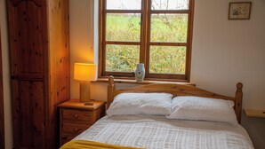 A bedroom with a wooden bed, a bedside table with a lamp, a window with a view of greenery, and a wooden wardrobe. - 3-bed Barn Eco-lodge on the Tarka Trail (Torrington)