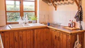 A kitchen with wooden cabinets, a sink, a window with a view of greenery, and a shelf with kitchenware. - 3-bed Barn Eco-lodge on the Tarka Trail (Torrington)
