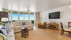 A spacious living room with a beige sofa, a wicker chair, a small table, and a television mounted on the wall.