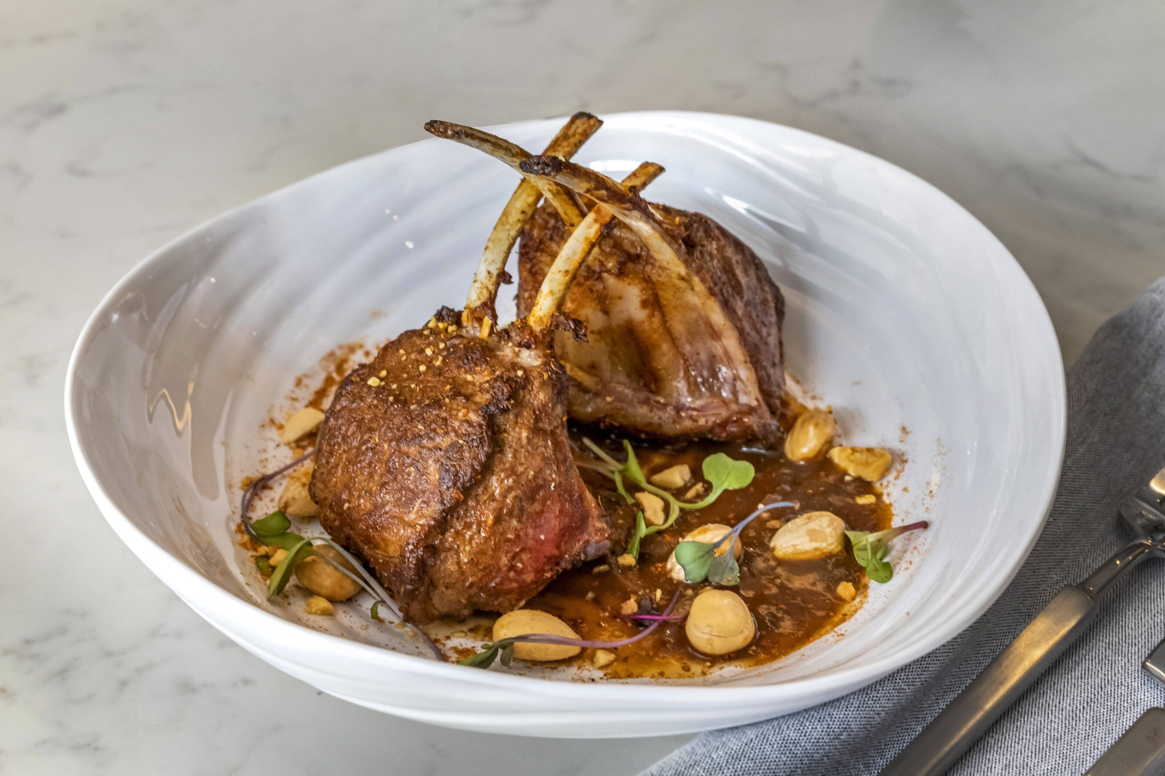 A white ceramic plate with two lamb chops, garnished with microgreens and peanuts, on a marble surface.