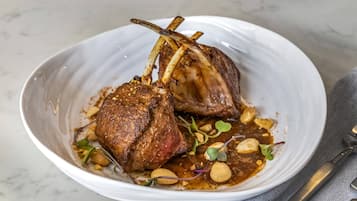 Un piatto di ceramica bianca con due costolette d'agnello, guarnite con microgreens e arachidi, su un ripiano di marmo.