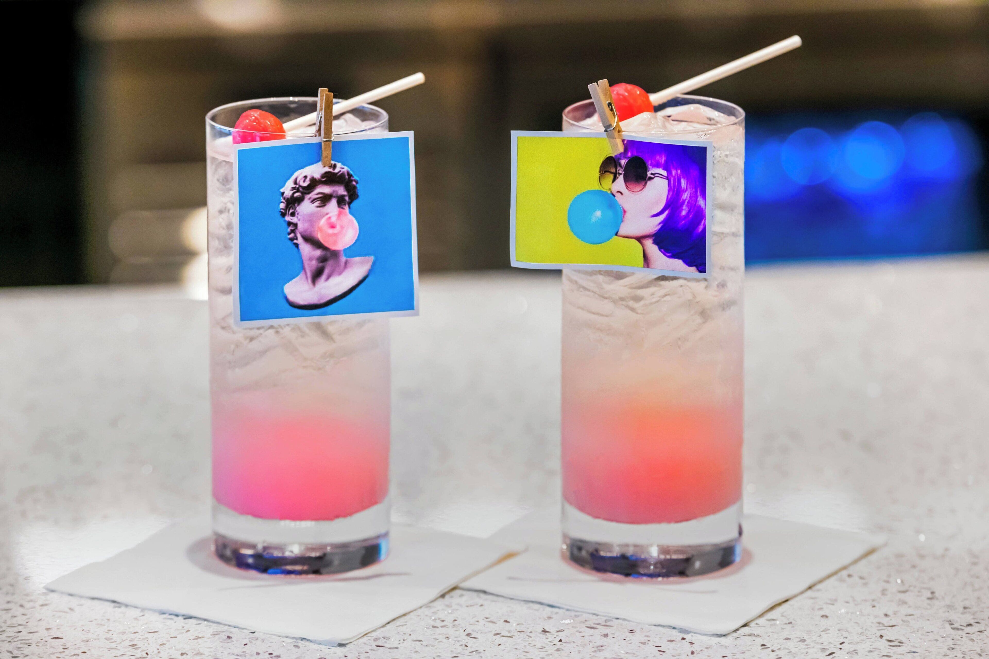 Deux cocktails aux couleurs vives, présentés en couches et décorés de petites images sur le bord du verre, sont posés sur une surface blanche.