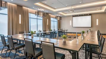 Un salón de conferencias con una larga mesa, sillas, una pantalla de proyector y ventanas amplias.