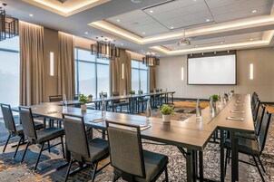 Un salón de conferencias con una larga mesa, sillas, una pantalla de proyector y ventanas amplias.
