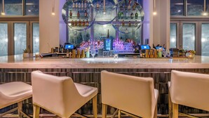 Zona de bar con diseño moderno, que incluye una barra central con una variedad de botellas y vasos, y asientos con taburetes altos.