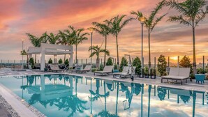 Zona junto a la piscina con sillones de descanso, una pérgola y palmeras al atardecer.