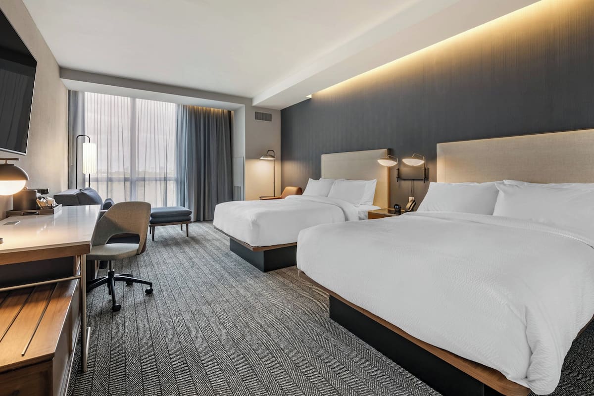 Une chambre d’hôtel avec deux lits, un bureau, une chaise et une grande fenêtre avec des rideaux.