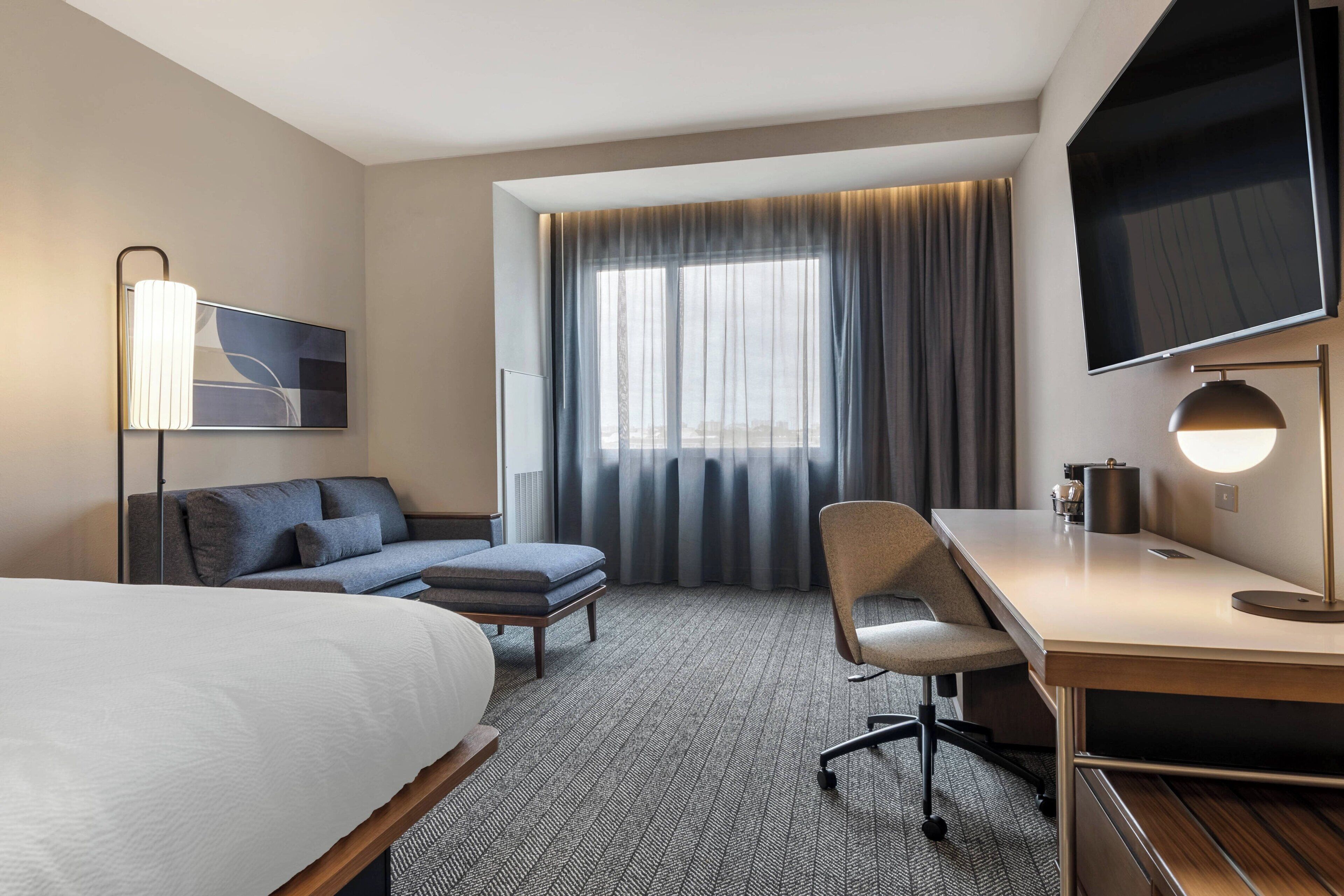 Habitación de hotel con cama, escritorio, silla, televisión y ventana con cortinas.