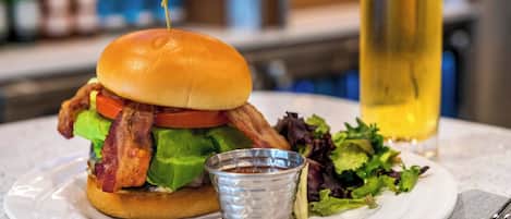 Een hamburger met bacon, sla en tomaat, een klein bakje saus en een glas bier op een bord.
