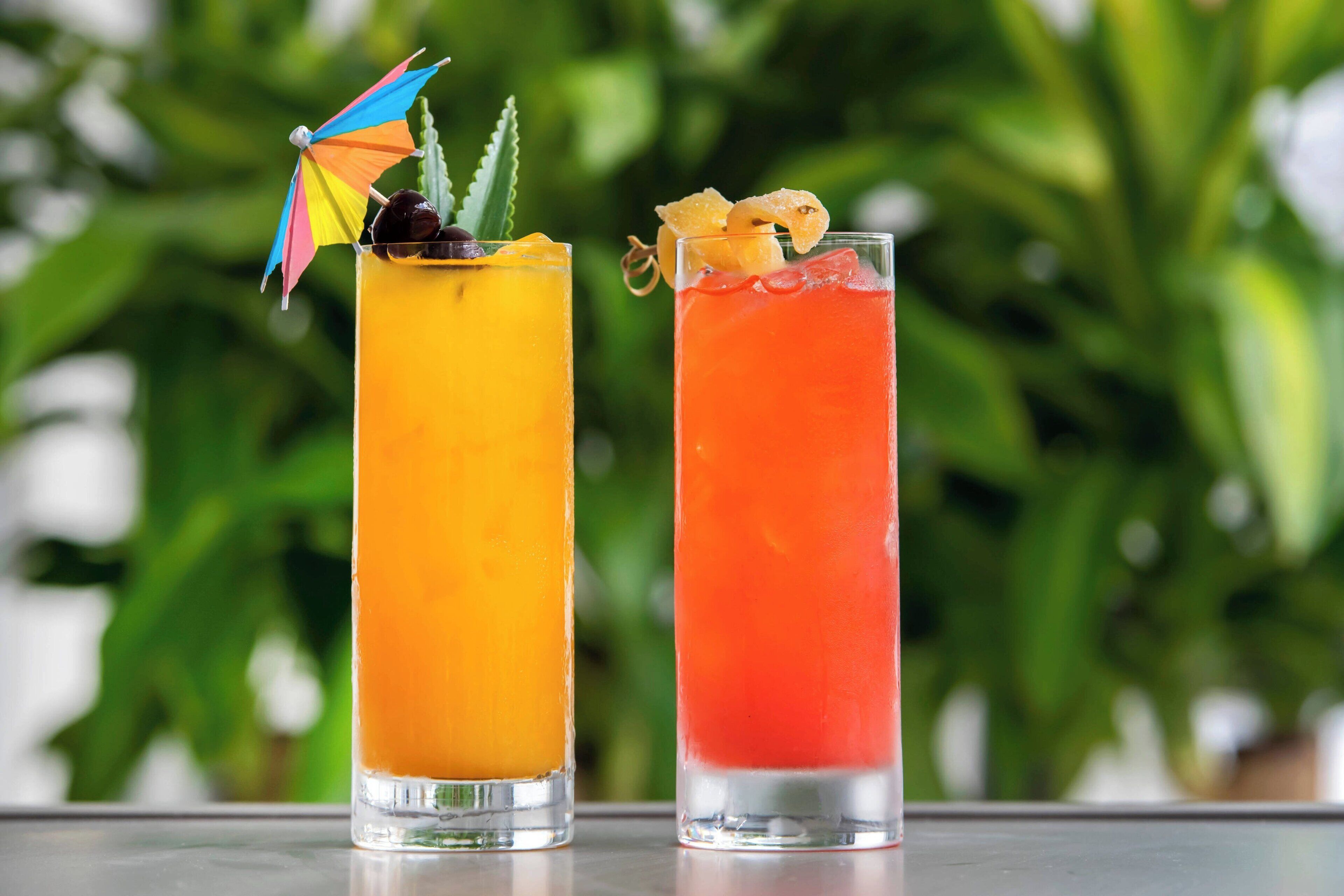 Deux grands verres de cocktails, décorés de fruits et d’autres éléments, posés sur une table devant un arrière-plan vert flou.