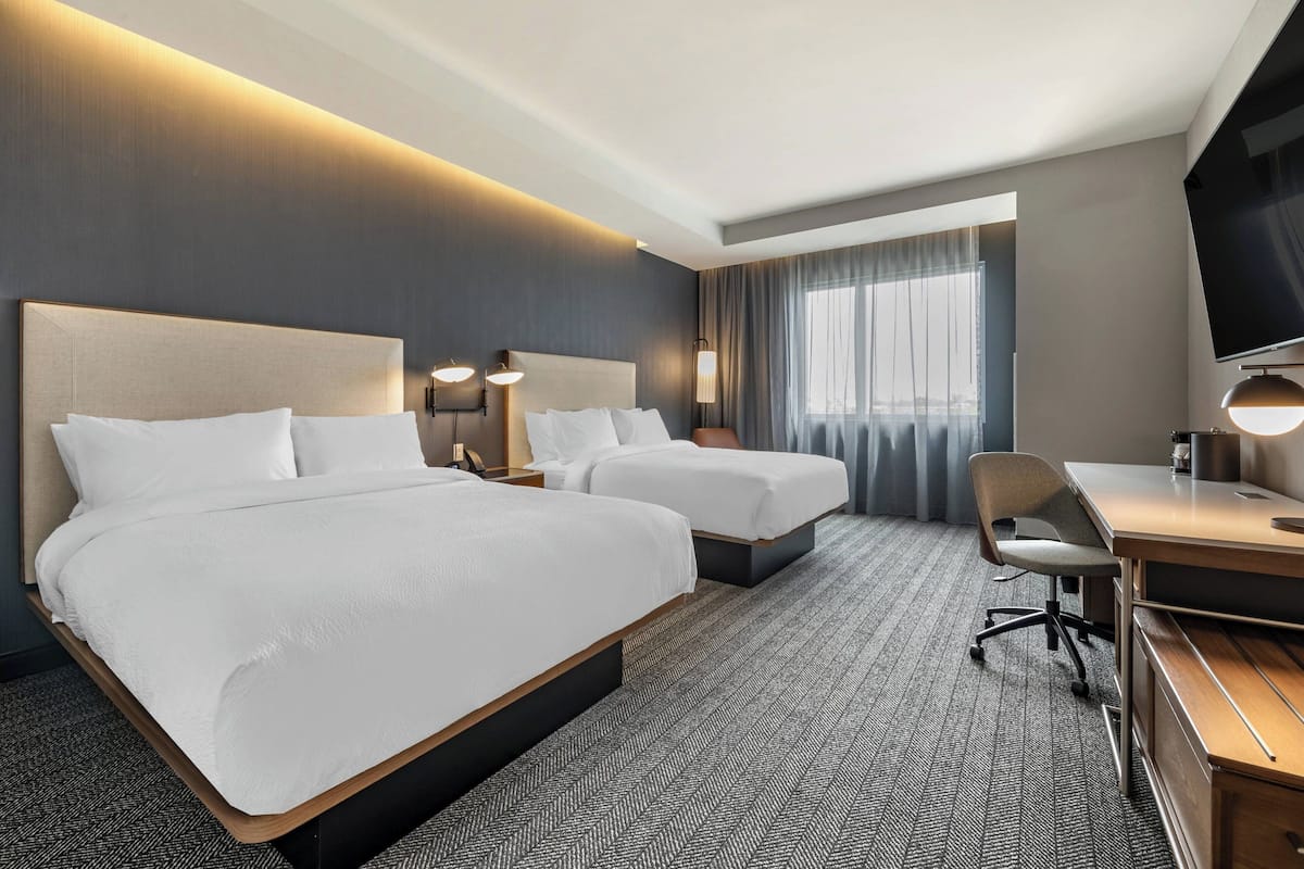 Une chambre d’hôtel avec deux lits, un bureau, une chaise, une télévision et une grande fenêtre avec des rideaux.