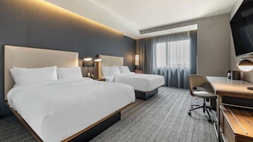 A hotel room with two beds, a desk, a chair, a TV, and a large window with curtains.