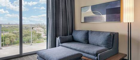 Une chambre d’hôtel moderne avec un canapé gris, un banc en bois, une lampe sur pied et une grande fenêtre donnant sur la ville.