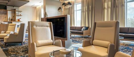 A modern hotel lobby with leather armchairs, a glass coffee table, a flat-screen TV, and decorative lighting.