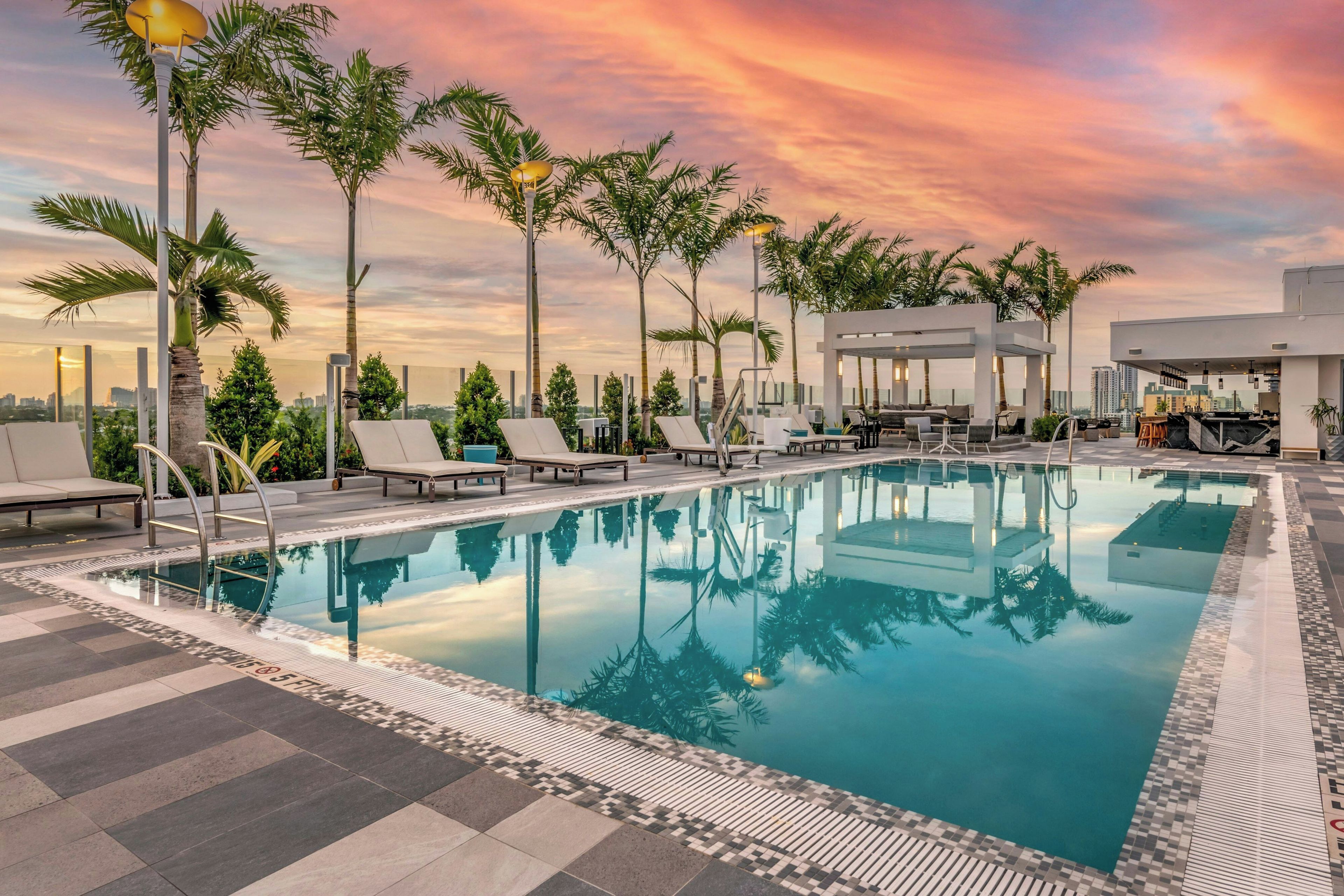A rooftop pool with lounge chairs and palm trees, surrounded by a cityscape.