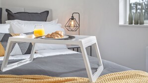 Una cama bien hecha con una bandeja de desayuno, una lámpara de noche y una ventana con botellas.