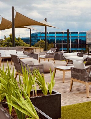 An outdoor seating area with wicker furniture and planters, overlooking a modern building with reflective glass windows. - Luxury Serviced Apartments Stevenage, Hertfordshire (Stevenage)