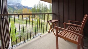 A balcony with a wooden table and two chairs, overlooking a mountainous landscape with trees and a road. - Incredible Views / Modern Updates / Hot Tub (Fernie)