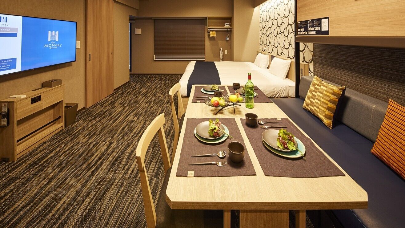 A hotel room with a dining area, a bed, a television, and a wall-mounted menu board.